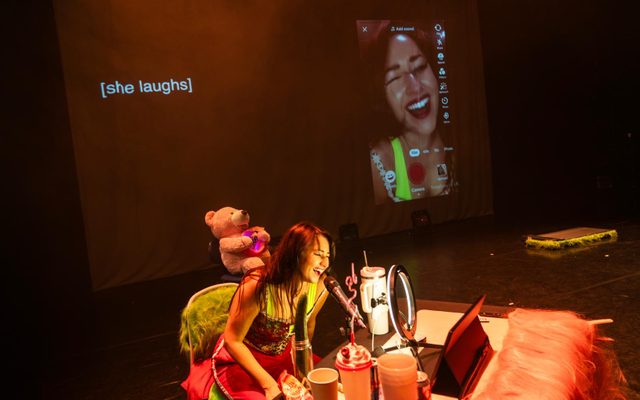 A picture of the set of the theatre production FAMEHUNGRY. Louise Orwin, a woman with black hair and green eyes, is sitting at a table laughing into a ring light, phone on a tripod, a microphone on her right and a large Stanley cup on her left. She is wearing bright pink tracksuit bottoms and a green and pink corset.