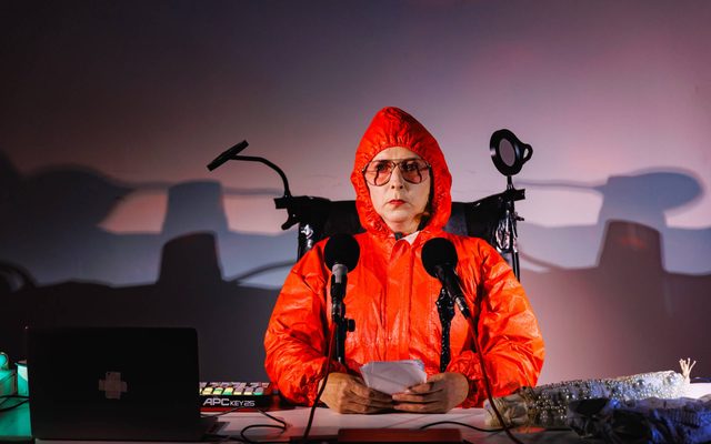A white woman with an emotionless expression is sitting at a desk behind two microphones; she is wearing a bright orange hazmat suite. Large, orange-tinted glasses and holding a piece of paper with both hand on the desk. On the table sits a laptop and electronic keyboard to the left, and two abstract bundles of fabric with pearl embezzlements.