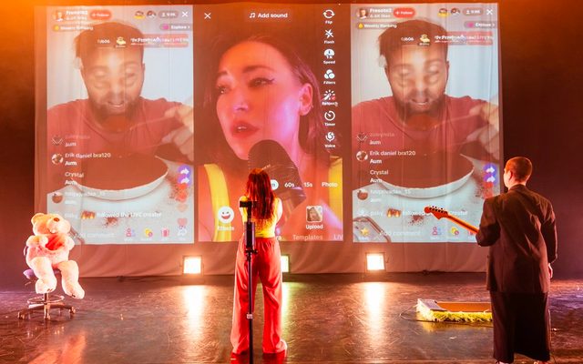 Louise Orwin stands in front of a screen projecting three phone screens on social media. In the middle is a livestream of her recording herself with her phone; either side is a person eating an enormous bowl of red jelly on a livestream, there are various likes and comments from fans on screen. Right of Louise is a giant teddy bear on an office chair, on on the left a person in all black holding a guitar.