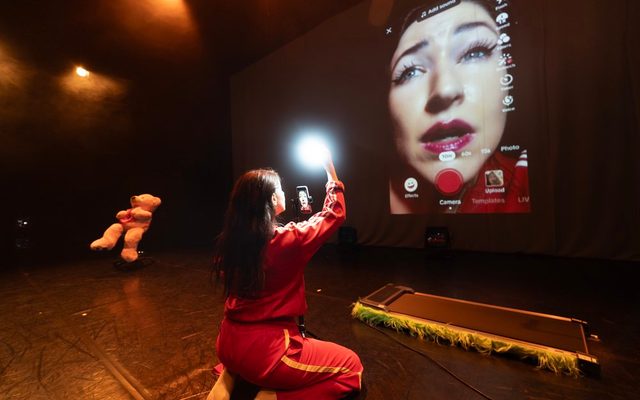 Actor Lousie Orwin kneels facing away from the camera, above her head she is holding a phone and flashlight being shone in her face; she is wearing a bright pink tracksuit and white socks. A livestream from the phone is being projected on the back wall behind her, and a large pink teddy bear sits on a chair in the corner.