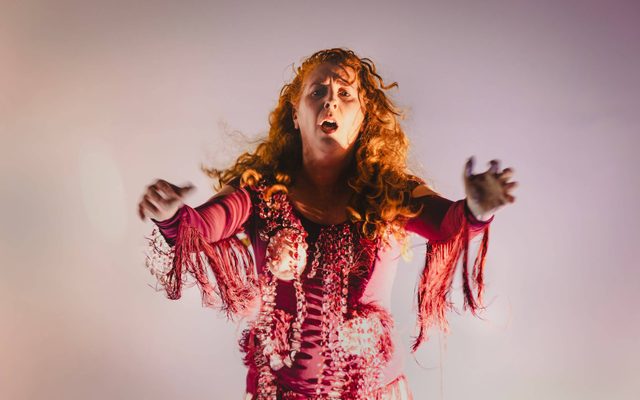 Catherine Hoffman, a white woman with long wavy ginger hair is standing face on to the camera with hands reaching forward and a somewhat confused zombie-like expression on her face. The background is white and grey, contrasting her colourful silhouette. She is wearing a dark pink long-sleeved leotard with strings of large light pink jewels and pink cloth tassels under the arms, swinging with movement.