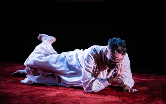 A man wearing an off-white-coloured full-body robe is laying on a red velvet carpet with his head closest to the camera. One hand is placed on the floor, the other reaching for his forehead.