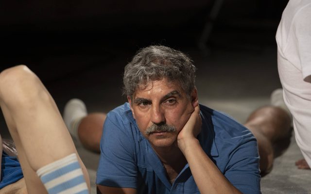 An older man with grey hair and a large moustache is lying on his front in a blue short-sleeved shirt. His face is propped up by one hand as he stares outwards. He’s surrounded by people who are mostly out of shot. There is one person in a white top whose back we can see, and another person’s leg is visible wearing a blue and white stripy sock.