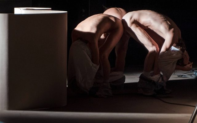 people are crouched over, facing outwards from each other with their bottoms together. They are mostly naked with their trousers at their ankles and their tops pulled over their heads. Next to them is a large piece of card that is freestanding, arranged in a curving s-shape running alongside them. 