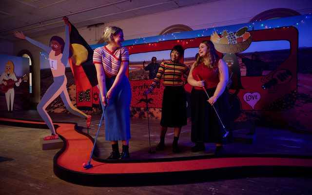 A vibrant min-golf installation with a winding red course with black boarders. A woman wearing a blue skirt and striped top is putting it along the track; she is looking over her shoulder and smiling at two other people. Behind them a large board is printed with a sandy desert and an animated big red bus. There is a cardboard cut-out of a runner with the label ‘1’ on their chest, they hold an Australian Aboriginal flag behind them.