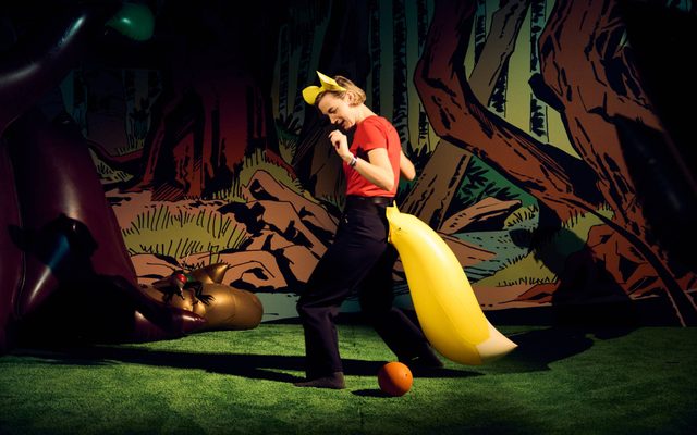 A person dressed up as a squirrel with ears and a large orange blow-up tail, is standing on fake grass next to a large orange ball, in front of a comic-style woodland backdrop. In the back left of the room there is a blow-up tree and fly.