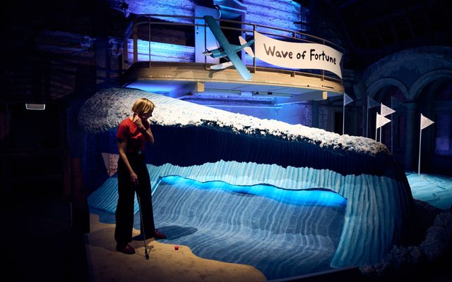 'Wave of Fortune’ min-golf installation for production Swingers, featuring a 6-foot wave made from plaster and lit with blue light, which must be put through on the course; a woman in a red top and black trousers is getting ready to put a ball. There are flagpoles on the other side of it and a seaplane above the scene.
