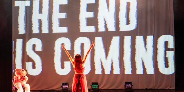 A woman wearing pink tracksuit bottoms and a crop top is standing facing away from the camera with arms held high and peace signing with her fingers. Behind her, a large screen reads ‘The End Is Coming’ in bold white distorted text on a black background. A large pink teddy bear sits on a chair in the corner.