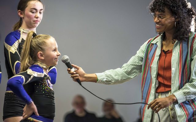 A Lady with dark brown curly hair and gold-rimmed glasses, wearing a horizontally striped green, red and blue shirt and a orange top, is interviewing two girls with a microphone outstretched to them. The girls are wearing black and blue sparkly leotards.