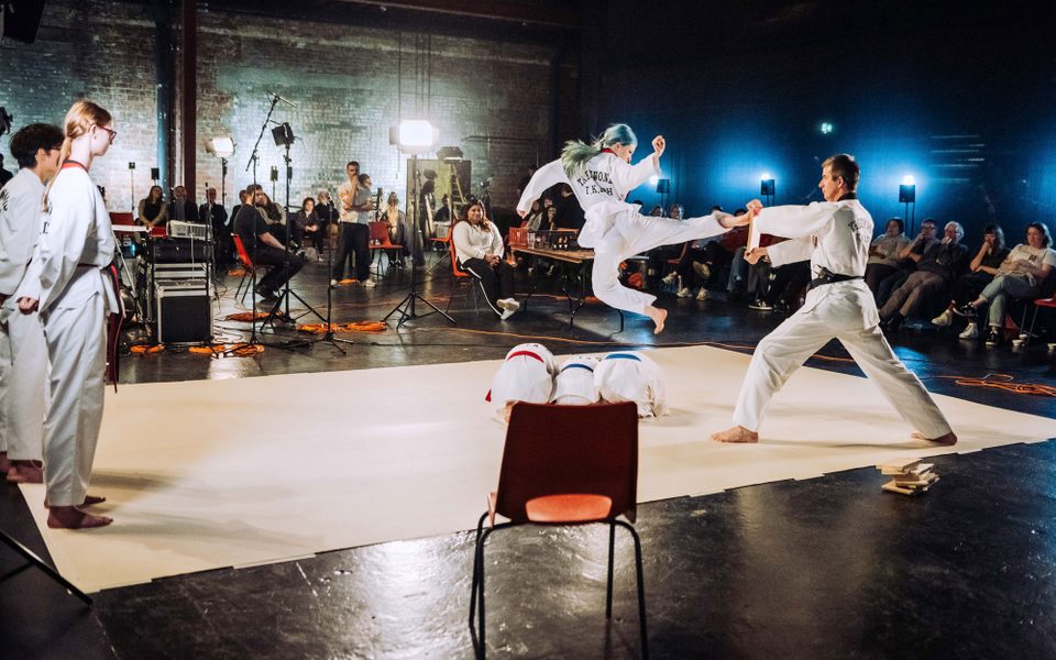 A karate showcase with a girl with blue hair wearing full Gi (white formal robes worn in various martial arts), who has jumped over three small hurdles and is fly-kicking a plank held by a man. In the background there is a curious audience and lots of sound equipment like microphones and trailing cables.
