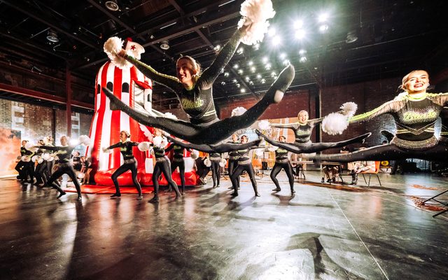 Dancers wearing black and yellow sparkled leotards are dancing in a large room with black polished flooring. The woman at the centre are in the height of a jump with legs outstretched into splits and arms up holding white pom poms. In the background there is a red and white striped blow-up house.
