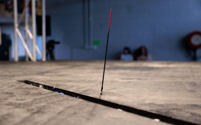 A black stick with a red tip stands upright in front of a rectangle slot cut out in the grey scratched stage.