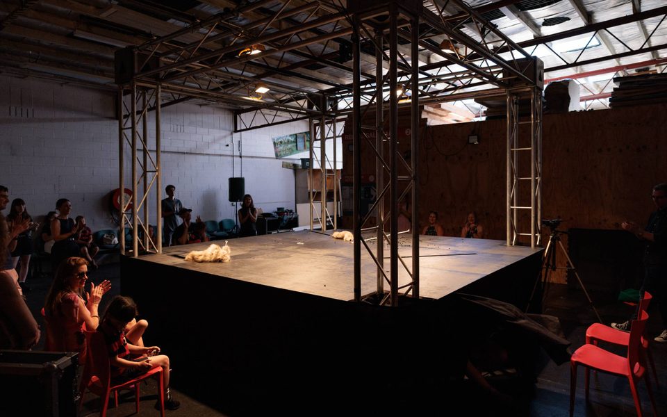 A small stage with metal pylons on each corner sits in the middle of a room, There is an audience sitting around it clapping.