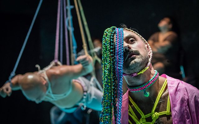 A person with techno-colour rope braids, necklace and chest rope tied in Shibari fashion, is gazing upward with a gentle sombre look on his face. Behind him a person is suspended in air with rope face down.