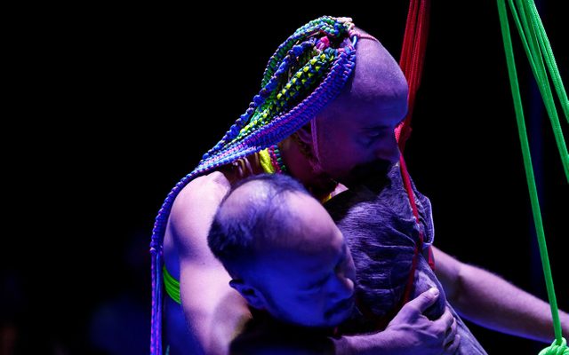 A person with techno-colour rope braids is standing behind another man, gently but firmly holding him to his chest, the other man is mostly out of shot, but he is suspended with neon red and green ropes that intersect and are woven around his body.