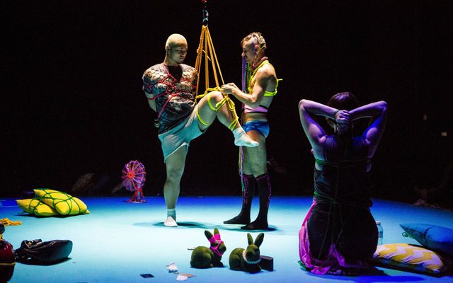 In the set of Bunny, various mundane objects like pillows and a desk fan lay on the light blue floor, tied in neon-coloured Shibari rope. In the centre one person is being tied up by the other, with one leg suspended in air, and the other on the floor, hands tied behind their back. They both gaze softly at the rope being tied, as another person with their back faced toward the camera, kneeling down fully woven in rope, watches the scene as well.