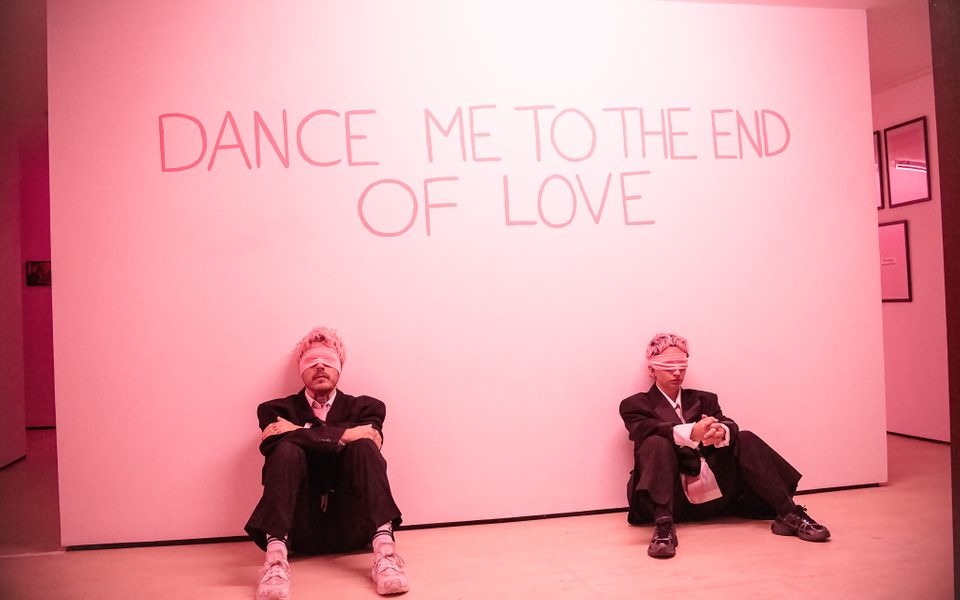 In a softly lit pink room the back wall reads 'dance me to the end of love' two people are sitting against the wall, wearing blindfolds, black suits and trainers.