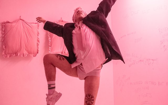 In a light pink lit room, a person stands dancing, they are wearing black blazer, white shirt and tie, with no trousers.