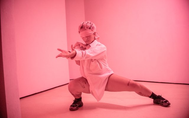In a light pink lit room, a person squats low to the ground, posing with their hands forward. They are wearing a blindfold, white shirt and tie and no trousers.
