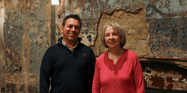Tarek Iskander is a brown-skinned man with dark curly hair wearing a three-quarter zip sweatshirt. He is smiling and stood next to Lyn Gardner, a white woman with a short grey bob who is wearing a pink cardigan. They are both standing inside in front of a wall that has distressed plaster and exposed brickwork.