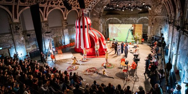A giant inflatable helter skelter in red and white candy-cane stripes takes up a large portion of BAC's Grand Hall, filled with an audience on one side looking on. There are gymnasts performing in gold leotards, twirling hula hoops and posing in the splits in fron of the slide. There is a screen at the back of the performance space with writing projected onto it. Lots of people are stood in the performance space staring at the helter skelter.