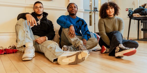 Three young people are sat on the floor od a studio space with light wood floor, staring proudly down the lens of the camera. They are of varying races and genders, and all are casually dressed.