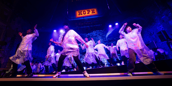 A group of young people dancing on stage. They are each wearing white t-shirts, black jeans, and a transparent poncho. There is a sing above them saying 'Hope'.