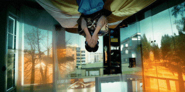 An image of a young person sitting on the edge of their bed with their hands covering their face. The young person and the bed are situated upside down as if stuck to the ceiling. The walls around the room have projections of buildings, trees and outdoor spaces.