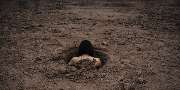 A young woman is submerged in a hole in an empty field of dirt. Only the back of their head and shoulders is visible.
