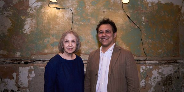 Two people stand against a worn painted wall. Left is Lyn Gardner. A woman with grey short hair with pale features wearing a navy long sleeve shirt. Right is Tarek Iskander. A man with dark hair wearing a tan blazer and white shirt.