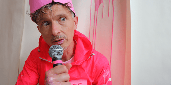 A white man with short grey hair is sat down and talking into a microphone. He’s wearing a pink paper crown on his head and a pink waterproof jacket with a sticky note on it that says ‘ME’. Behind him a pale pink liquid is dripping down the wall. The same liquid is also on his jacket, hand and lips as well.