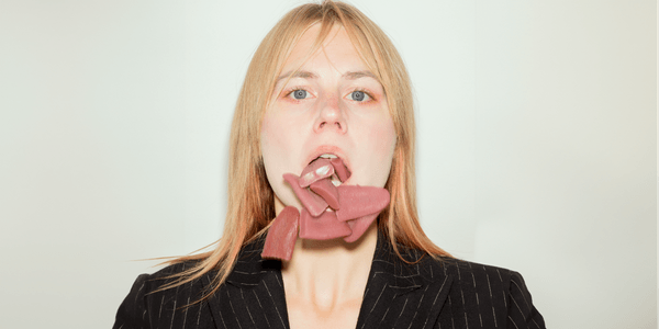 A headshot of a white woman with blonde hair and blue eyes in a black and white pinstriped suit. Her mouth is open, and lots of tongues are falling out of it.
