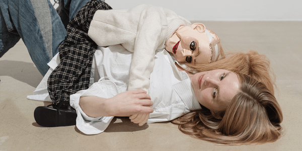 A white woman with blonde hair is lying on her back, cuddling a ventriloquist doll. She’s wearing a white shirt and jeans, and the doll is also blonde, with lined eyes and very red lips. It’s wearing a cream turtleneck jumper and chequered black and white trousers.