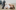 A white woman with blonde hair is lying on her back, cuddling a ventriloquist doll. She’s wearing a white shirt and jeans, and the doll is also blonde, with lined eyes and very red lips. It’s wearing a cream turtleneck jumper and chequered black and white trousers.