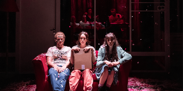 Three people are sat on a red sofa on a stage, surrounded by red and pink confetti. There is one man on the left-hand side in jeans and a t-shirt, he has blonde hair in a ponytail and green paint in war-like markings across his face. The other two are women – one on the right-hand side of the sofa is wearing a green hoodie and combat boots, and the one in the centre is wearing orange joggers and is holding a cardboard box.