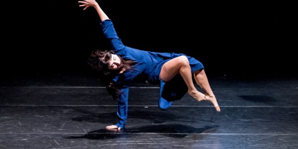 A woman with dark hair wearing a blue dress suspends herself from the floor of a dark stage with one arm, the other is raised above her and her legs are raised off the floor.