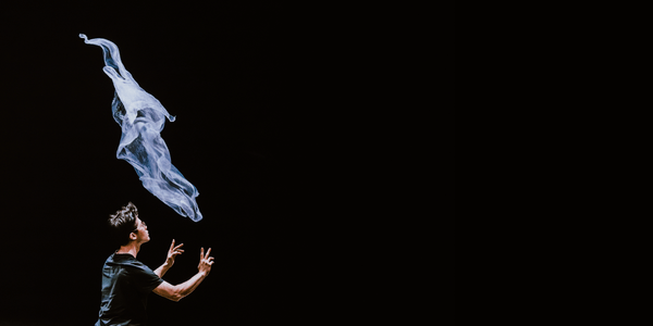 A man with short black hair and glasses is has their back slightly turned away from the viewer, wearing a black t-shirt. He has just thrown a pale, almost transparent piece of cloth in the air, his arms still outstretched from the motion. The cloth is still suspended in the air. The background is completely black.