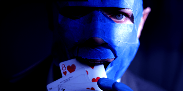 A white man with piercing blue eyes is mostly in shadow, with only the right edge of his face in the light. He has blue tape all over his face, covering everything but his eyes, nostrils and mouth. He is holding some playing cards to his mouth as if he is about to eat them, his hand is in a blue latex medical glove.