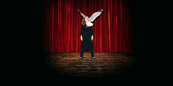 A woman is stood on a theatre stage in front of a red curtain. She’s white, wearing all black and has long brown hair. A seagull is flying towards her, blocking us from seeing her face.