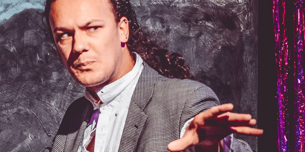 A white man with long curly hair tied back in a ponytail is wearing a suit and extending out his arms as if ready to cast a spell. He looks concerned, as if he’s trying to ward something off. Behind him is a window painted black and a metallic red and purple curtain. The whole image has a paint-like quality to it, as if it’s been put through a retro filter on photo-booth to make it appear more like a poster.