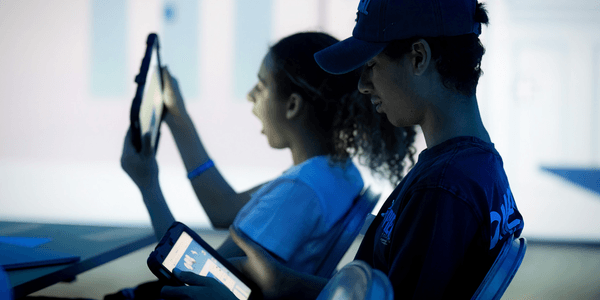 Two young people, a boy and a girl, are holding iPads in a dark room with a bright projected screen on the wall behind them. The boy is wearing a t-shirt and cap, and is looking thoughtfully at his iPad which is also illuminating his face. The girl is holding the iPad up close to her face and tilting it slightly, her mouth open as she smiles at what she sees.