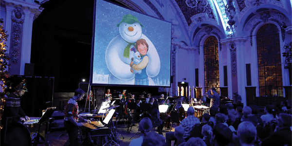Live orchestra performing The Snowman and the Snowdog beneath a large screen in an ornate, blue-lit concert venue
